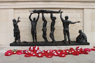 A group of bronze sculptures depicting soldiers hoisting a wounded comrade on a stretcher against the backdrop of a memorial wall. A woman and child stand nearby, while another soldier kneels on the ground. Bright red poppy wreaths are arranged in front of the memorial.