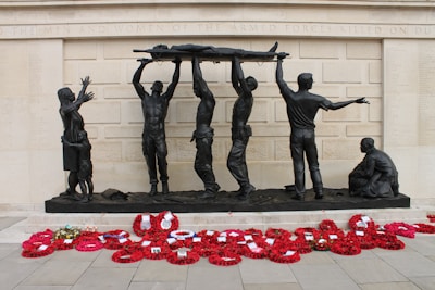 A group of bronze sculptures depicting soldiers hoisting a wounded comrade on a stretcher against the backdrop of a memorial wall. A woman and child stand nearby, while another soldier kneels on the ground. Bright red poppy wreaths are arranged in front of the memorial.