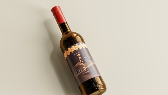 A wine bottle with a modern label design, featuring a red bottle cap and an elegant patterned label. The bottle rests on a light-colored surface, casting a soft shadow.