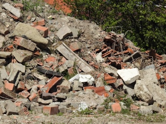 A pile of construction rubble consisting of broken bricks, concrete blocks, and other debris. The rubble is scattered across the ground with some greenery interspersed among the debris. The background shows vegetation with some bushes or small trees.