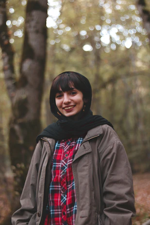 A smiling person wrapped in a warm shawl, holding a Calmaraiz product while sitting outdoors among trees.