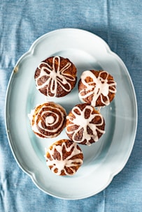 Five pastries with white icing designs are arranged on a light blue oval plate, which is placed on a blue fabric background.