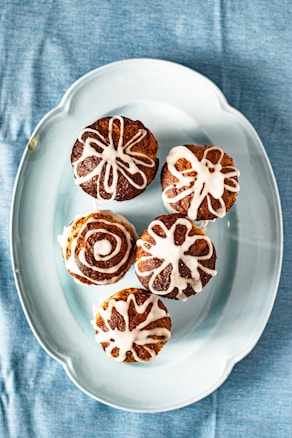 Five pastries with white icing designs are arranged on a light blue oval plate, which is placed on a blue fabric background.