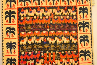 A vibrant textile featuring rows of stylized animal and human figures in a colorful tapestry. The motifs include elephants, camels, and human figures, highlighted by decorative patterns such as flowers and geometric shapes. The border contains repetitive tree and human motifs, enhancing the overall aesthetic.