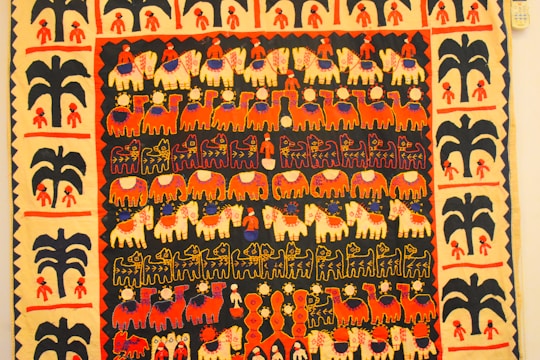 A vibrant textile featuring rows of stylized animal and human figures in a colorful tapestry. The motifs include elephants, camels, and human figures, highlighted by decorative patterns such as flowers and geometric shapes. The border contains repetitive tree and human motifs, enhancing the overall aesthetic.