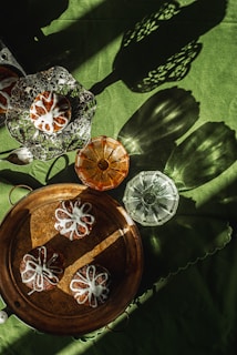 A collection of glass dishes and pastries with white icing drizzled in a floral pattern are displayed on a green surface. There are intricate dishes, including a multi-tiered platter holding pastries, a large round tray with pastries, and two ornate glass coasters, one amber and one clear.