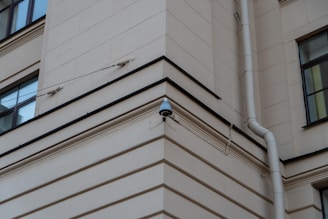 A security camera mounted on the corner of a beige building, with a drainpipe running vertically along the wall. The building features square windows and simple architectural lines.