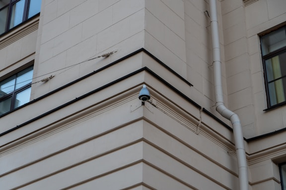 A security camera mounted on the corner of a beige building, with a drainpipe running vertically along the wall. The building features square windows and simple architectural lines.