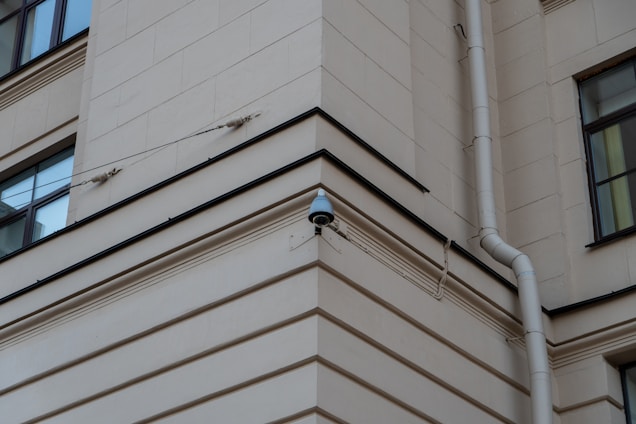 A security camera mounted on the corner of a beige building, with a drainpipe running vertically along the wall. The building features square windows and simple architectural lines.