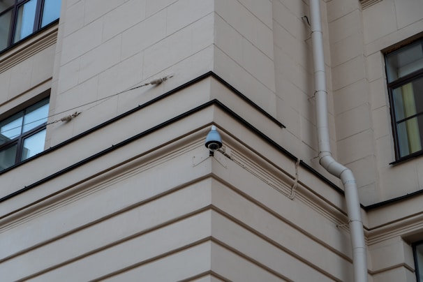 A security camera mounted on the corner of a beige building, with a drainpipe running vertically along the wall. The building features square windows and simple architectural lines.