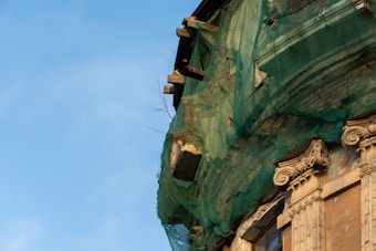 An old, partially crumbling building with green mesh netting draped over its facade is visible. The architectural elements include detailed columns with ornate capitals. The building appears to be under renovation or preservation efforts.
