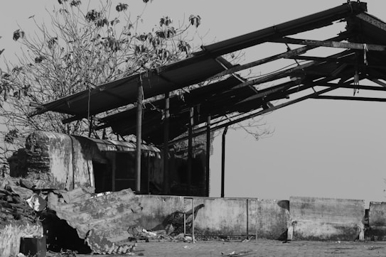 A black and white photo of a dilapidated structure with a partially collapsed roof. The building is surrounded by debris, including corrugated metal sheets. In the background, bare trees are visible, adding to the scene's sense of abandonment.
