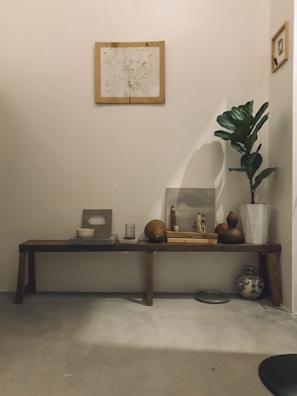 A minimalist interior with a wooden bench against a neutral wall displays an arrangement of decorative items. Above hangs a framed abstract artwork. On the bench are geometric pottery pieces, round gourds, a candle, and a small statue. A potted plant with wide green leaves stands to the right, next to an ornate vase on the floor.