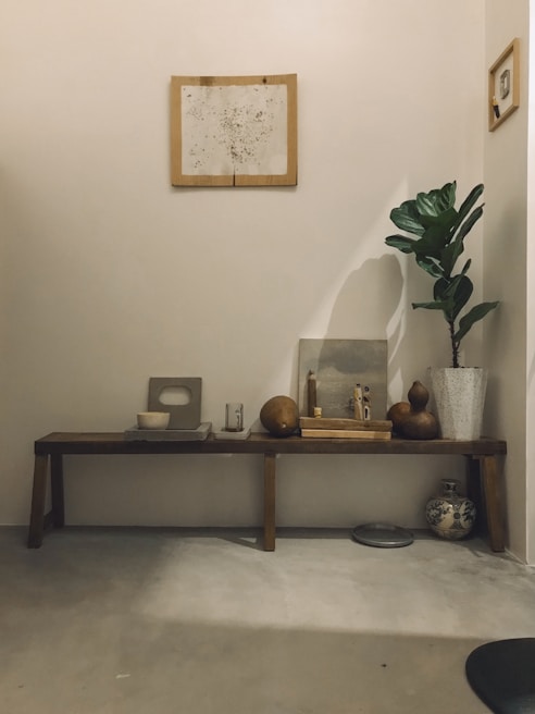 A minimalist interior with a wooden bench against a neutral wall displays an arrangement of decorative items. Above hangs a framed abstract artwork. On the bench are geometric pottery pieces, round gourds, a candle, and a small statue. A potted plant with wide green leaves stands to the right, next to an ornate vase on the floor.