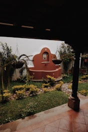 A serene garden courtyard features a red terracotta-colored fountain against a white wall, adorned with a small religious icon in a niche above it. Two hanging plants with cascading fronds frame the scene, and well-maintained green grass is bordered by a cobblestone path and manicured shrubs. The shaded porch area with a tiled floor and wooden post provides a tranquil, sheltered space.