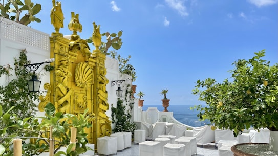 A rooftop terrace overlooking the sea with vibrant yellow architectural features and decorative sculptures. The terrace is adorned with potted plants such as lemon trees and other greenery. White cube-shaped seats are arranged around the space, and the sky is clear with a few clouds.