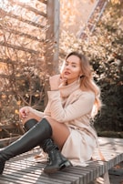 A young woman wearing a cozy sweater and boots, sitting on a bench with autumn leaves around.