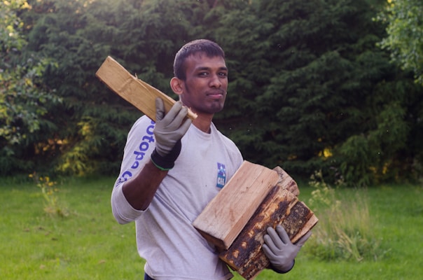 A person wearing gloves is holding a few pieces of chopped firewood outdoors. The background consists of lush green shrubs and grass, suggesting a garden or forest setting.