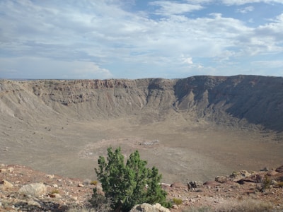 A vast, circular meteor crater with steep rocky walls and a barren, dusty interior. The sky is partly cloudy, providing a backdrop to the dramatic landscape. Vegetation is sparse, with a few small green shrubs visible at the crater's edge.