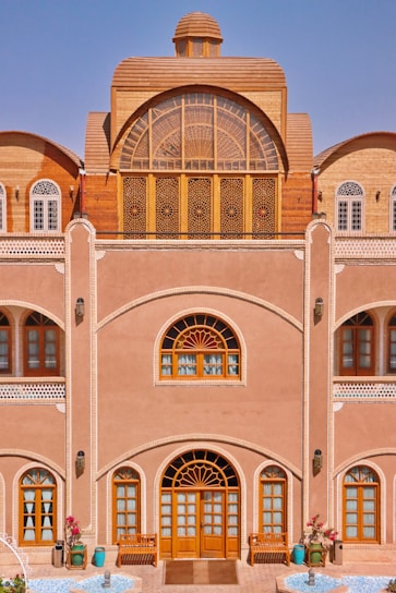 A traditional building with intricate wooden windows and doors, an arched design, and detailed latticework. The façade features decorative elements with a natural earthy tone, complemented by blue sky in the background. There are potted plants and a small water feature at the entrance, adding a touch of nature to the architecture.