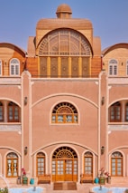 A traditional building with intricate wooden windows and doors, an arched design, and detailed latticework. The fa&ccedil;ade features decorative elements with a natural earthy tone, complemented by blue sky in the background. There are potted plants and a small water feature at the entrance, adding a touch of nature to the architecture.
