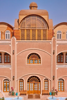 A traditional building with intricate wooden windows and doors, an arched design, and detailed latticework. The façade features decorative elements with a natural earthy tone, complemented by blue sky in the background. There are potted plants and a small water feature at the entrance, adding a touch of nature to the architecture.