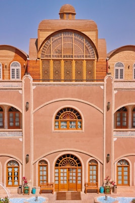 A traditional building with intricate wooden windows and doors, an arched design, and detailed latticework. The fa&ccedil;ade features decorative elements with a natural earthy tone, complemented by blue sky in the background. There are potted plants and a small water feature at the entrance, adding a touch of nature to the architecture.