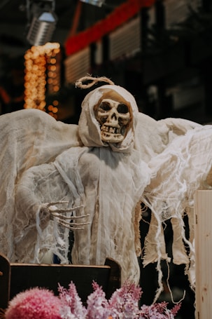 A grim reaper figure with a skull wearing tattered white cloth, positioned in a setting with blurry background lights and some pink flowers in the foreground, creating a spooky ambiance.