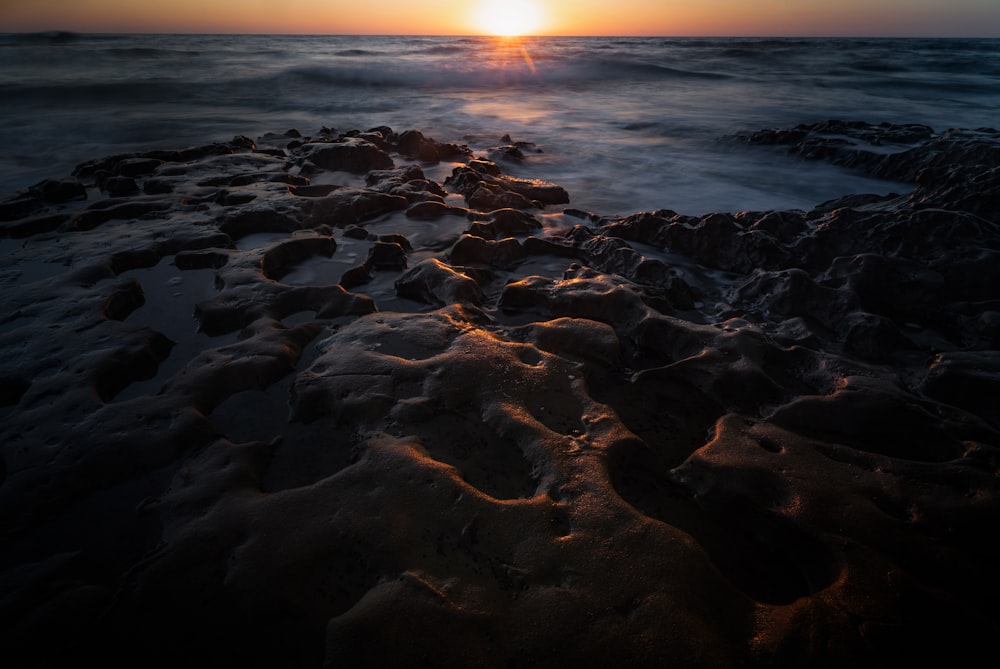 the sun is setting over the ocean on a rocky beach