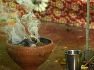 A clay incense holder emits swirling smoke against a richly patterned red and yellow floral background. A silver metal cup is placed nearby on the surface, which is scattered with small flower petals and remnants, evoking a sense of ritual or ceremony.