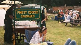 A food cart with a sign reading 'Finest Cooked Sausages' is situated in a grassy outdoor area. In the foreground, a woman is seated on a chair, appearing relaxed. Several people are gathered in the background, seated around picnic tables, suggesting a small outdoor gathering or event.