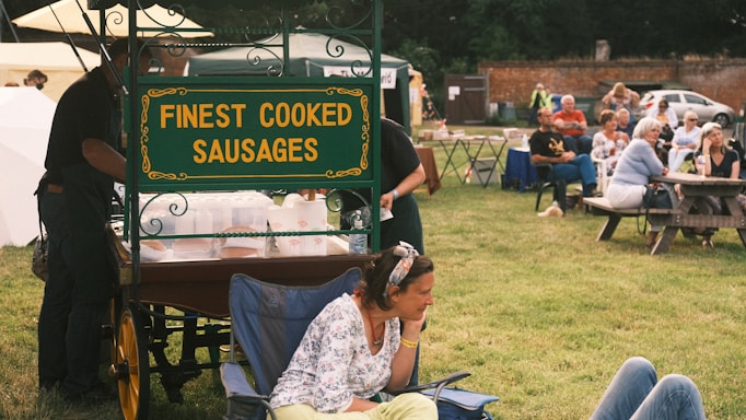 A food cart with a sign reading 'Finest Cooked Sausages' is situated in a grassy outdoor area. In the foreground, a woman is seated on a chair, appearing relaxed. Several people are gathered in the background, seated around picnic tables, suggesting a small outdoor gathering or event.
