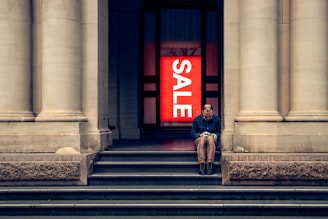 a man sitting on the steps of a building with a sale sign in the background