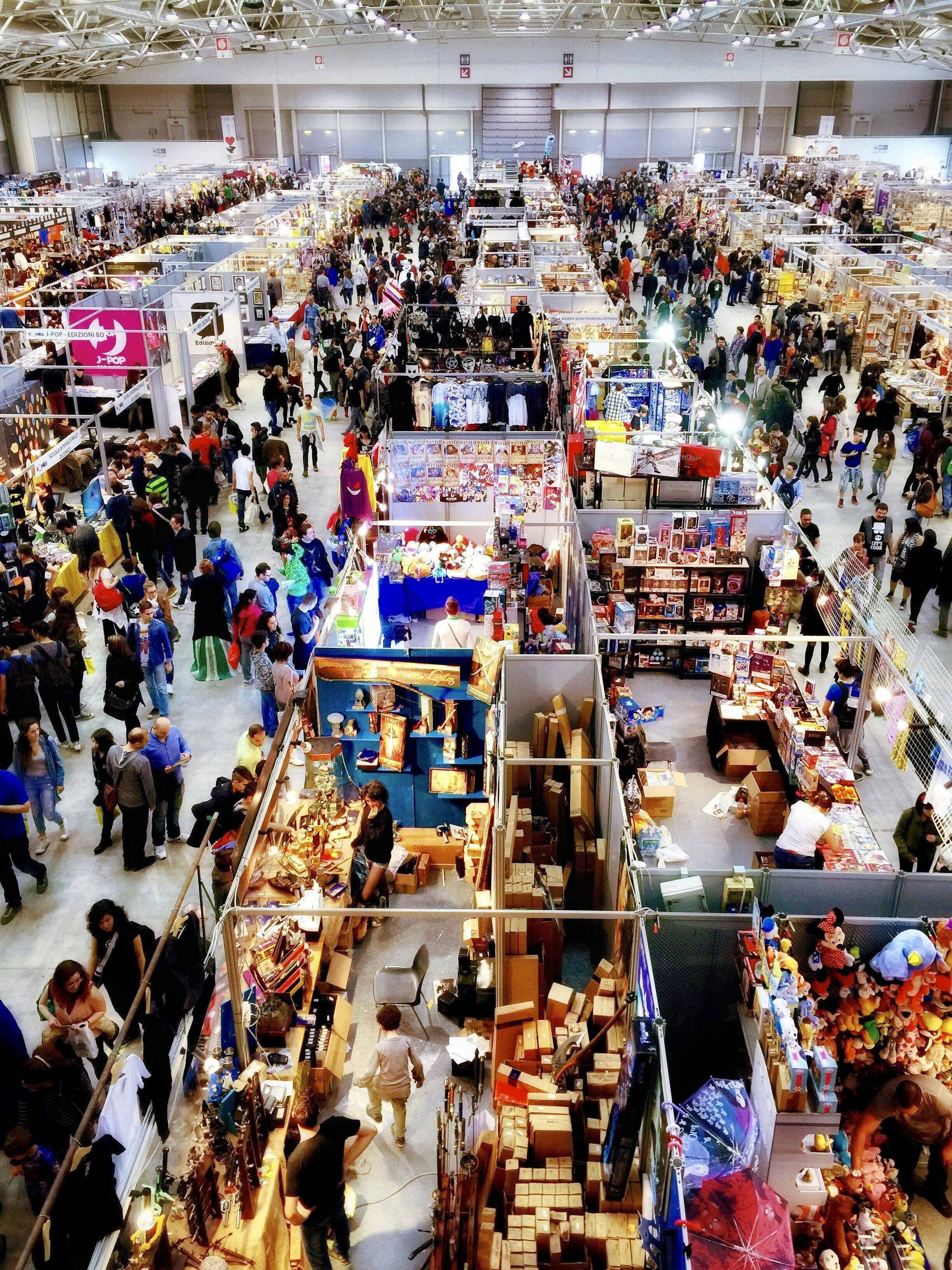 A crowded indoor market or convention center with numerous stalls selling a variety of items. People walk through the aisles, browsing merchandise and engaging with vendors. Colorful displays and products are visible, contributing to a lively atmosphere.