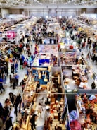 A crowded indoor market or convention center with numerous stalls selling a variety of items. People walk through the aisles, browsing merchandise and engaging with vendors. Colorful displays and products are visible, contributing to a lively atmosphere.