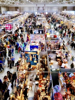 A crowded indoor market or convention center with numerous stalls selling a variety of items. People walk through the aisles, browsing merchandise and engaging with vendors. Colorful displays and products are visible, contributing to a lively atmosphere.