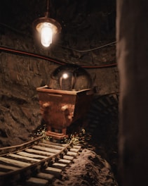 A dimly lit underground tunnel with a rusty mining cart on old wooden tracks. Overhead, a single bulb casts a warm, orange glow on the stone walls and cart. The scene includes sparking metal, creating a dynamic and gritty ambience.