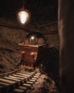 A dimly lit underground tunnel with a rusty mining cart on old wooden tracks. Overhead, a single bulb casts a warm, orange glow on the stone walls and cart. The scene includes sparking metal, creating a dynamic and gritty ambience.