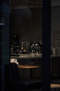 A dimly lit cafe interior with a counter featuring various coffee-making equipment. The space is primarily empty, with a single wooden table and padded seating visible in the foreground. A sign above the counter reads 'Mobile Order Pick Up.' The overall ambiance is subdued due to the low lighting.