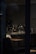 A dimly lit cafe interior with a counter featuring various coffee-making equipment. The space is primarily empty, with a single wooden table and padded seating visible in the foreground. A sign above the counter reads 'Mobile Order Pick Up.' The overall ambiance is subdued due to the low lighting.
