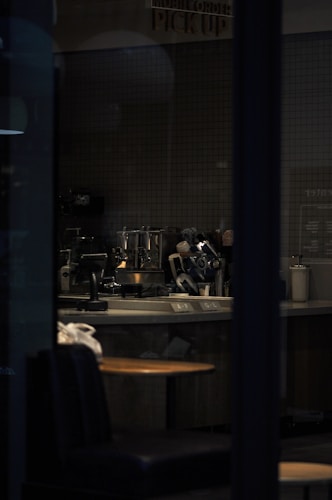 A dimly lit cafe interior with a counter featuring various coffee-making equipment. The space is primarily empty, with a single wooden table and padded seating visible in the foreground. A sign above the counter reads 'Mobile Order Pick Up.' The overall ambiance is subdued due to the low lighting.