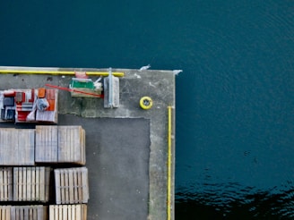 An aerial view of a concrete dock or pier extending into dark blue water. On the dock are stacked wooden pallets and various containers, next to a small green structure and a red barrier. Yellow lines and markings are visible on the dock.