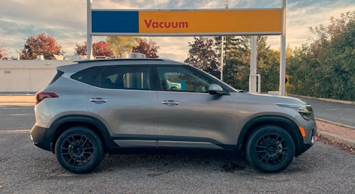 A silver SUV is parked in front of a vacuum station at a car wash. The background shows trees with autumn foliage and a partly cloudy sky. The vacuum station has a sign with red text on a blue and yellow background.