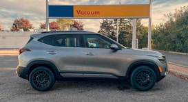 A silver SUV is parked in front of a vacuum station at a car wash. The background shows trees with autumn foliage and a partly cloudy sky. The vacuum station has a sign with red text on a blue and yellow background.