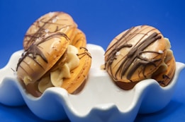 Three macarons filled with cream and chocolate ganache are placed in a white, wavy-edged dish. The macarons are drizzled with dark chocolate and sprinkled with coarse salt. The background is a vivid blue, which enhances the presentation of the macarons.