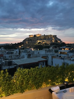 A scenic view of the Acropolis in Athens, Greece, with the ancient ruins illuminated against a dramatic evening sky. The skyline features urban buildings in the foreground and a gathering of dark clouds with hints of sunset colors in the background. A small dining area with a white tablecloth, plates, cutlery, and a candle is visible at the bottom.