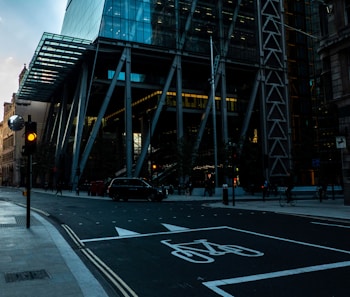 A street scene in an urban area features a modern glass and steel building. The structure has a prominent grid pattern and is supported by large metal beams. A traffic light is displayed with a yellow signal, and a black car is seen parked on the side. Bicycle lane markings are visible on the road, and a few pedestrians and cyclists are present.