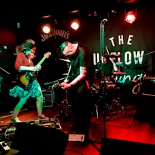 Musicians are energetically performing on stage with guitars, surrounded by amplifiers and a drum set. Bright stage lights illuminate the performance area, and a branded backdrop is visible behind the band.