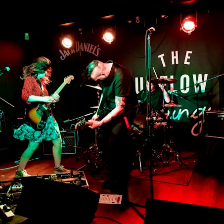 Musicians are energetically performing on stage with guitars, surrounded by amplifiers and a drum set. Bright stage lights illuminate the performance area, and a branded backdrop is visible behind the band.