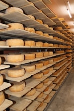 Rows of cheese wheels are neatly arranged on wooden shelves inside a storage or aging room. The shelves are stacked from floor to ceiling, and the room is well-lit with fluorescent lights.